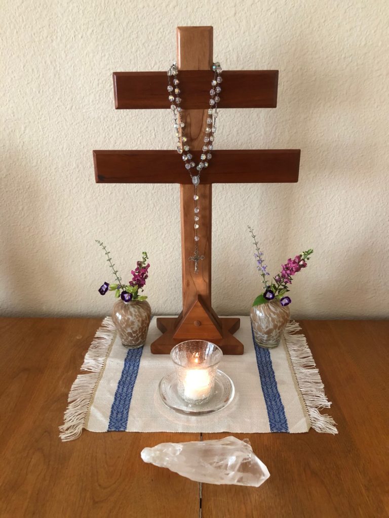 Santo Daime alter. A rosary sits on a cross, sitting upon a white and blue fabric place setting. On the place setting are two flower vases with flowers and in the centre of the setting is a lit candle. In front of the display is a large clear crystal.