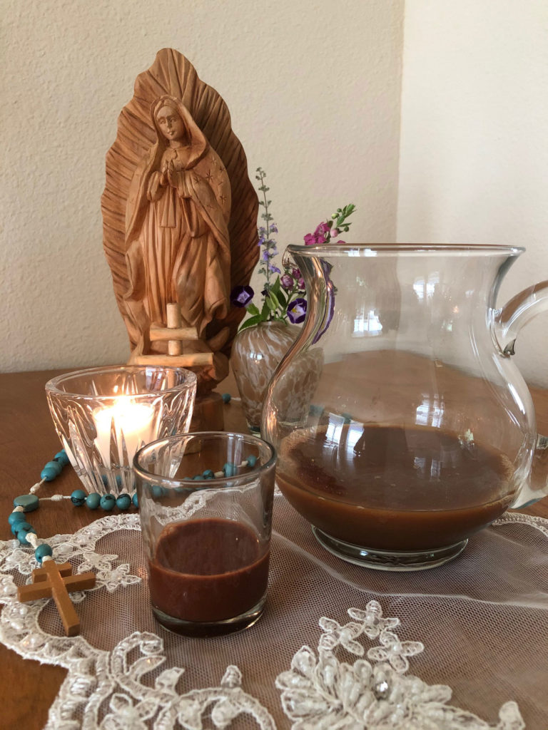 A Santo Daime alter consisting of a wooden sculpture of Mother Mary, a wooden cross, and a blue rosary. In front are a glass pitcher and glass, half filled with a Ayahuasca, a brown liquid.